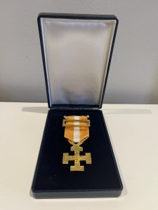 A gold cross medal with a gold pin attached to an orange and white striped medal ribbon with the Scouts logo engraved into the cross.