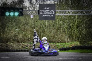 Henry racing over the finish line holding a chequered flag , and a sign above him saying 'Henry Greenwood All Star Champion'. 